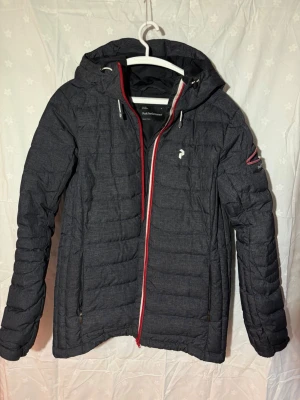 Mörkgrå dunjacka Peak Performance XL - Snygg mörkgrå dunjacka från Peak Performance i storlek XL. Jackan har huva, röd dragkedja och diskret logga på bröstet. Perfekt för kalla vinterdagar med sin quiltade design och värmande material. Snölås och skidkort ficka.
