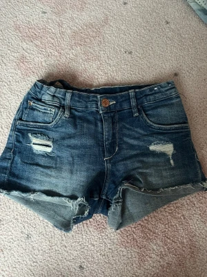Blå denimshorts med slitningar - Snygga blå denimshorts från H&M med slitna detaljer och fransig kant. Klassisk femficksmodell med knapp och dragkedja. Perfekta för en avslappnad stil under varma dagar.