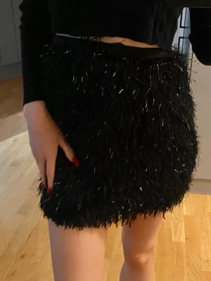Svart glittrig kortkjol med fransar - Snygg svart kortkjol med glittriga detaljer och fransar som ger en festlig look. Kjolen har en rak passform och är perfekt för dig som vill sticka ut. Passar till crop tops och klackar för en trendig outfit.