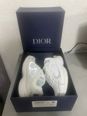 Vita D sneakers med mesh och läder - Säljer ett par vita D sneakers med chunky sula, detaljer i mesh och läder samt silverfärgade inslag. Skorna har en modern och sportig design med tydliga D-loggor på sidan. Perfekta för dig som vill ha en clean och exklusiv look.