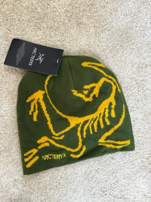Grön mössa från Arc'teryx med gul logga - Snygg grön mössa från Arc'teryx med stort gult fossiltryck och logga framtill. Tillverkad i mjukt stickat material som sitter skönt på huvudet. Perfekt för dig som gillar friluftsliv och vill ha en cool accessoar med tydlig profil.