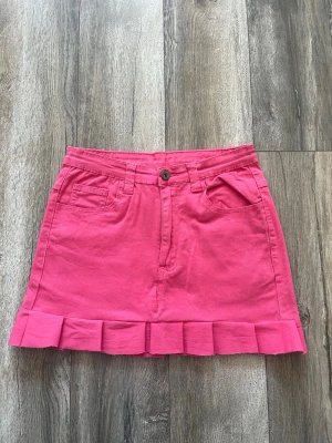 Rosa kjol med volangkant - Snygg rosa kjol i bomull med volangdetalj längst ner. Kjolen har fickor fram och bak, bälteshällor och stängs med knapp och dragkedja. Perfekt för dig som vill ha en färgglad och trendig look.