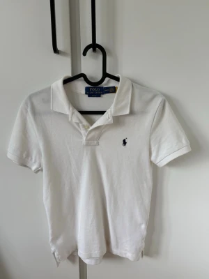 Vit pikétröja från Polo Ralph Lauren - Klassisk vit pikétröja från Polo Ralph Lauren med blå broderad logga på bröstet. Slim fit-modell med korta ärmar och krage med knappar. Perfekt för en clean och stilren look. Köpt på nk i Stockholm.