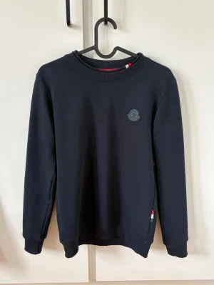 Svart sweatshirt från Moncler - Stilren svart sweatshirt från Moncler med rund halsringning och diskret Moncler-logga på bröstet. Tröjan har ribbade muddar vid ärmslut och nederkant samt en liten röd, vit och blå detalj vid sidan. Perfekt för en clean och avslappnad look.