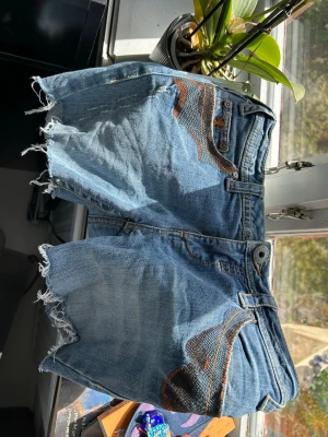 Blå jeansshorts med fransar och broderi - Säljer ett par blå jeansshorts från True esprit med råa fransade kanter och snygga broderade detaljer på fickorna. Klassisk femficksmodell med knappstängning och coola kontrastsömmar. Perfekta för en avslappnad och trendig look.