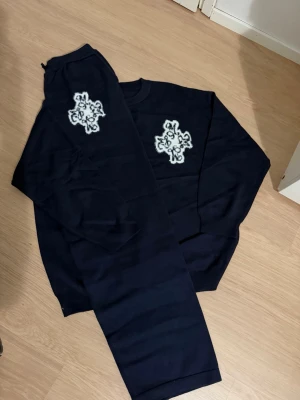 Mörkblått loungewear-set med brodyr - Mysigt mörkblått loungewear-set med matchande tröja och byxor. Båda plaggen har en vit broderad detalj framtill och på benet. Setet har en avslappnad passform och är perfekt för hemmadagar. Materialet ser ut att vara mjukt och bekvämt.