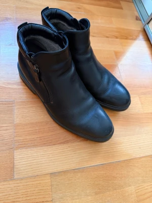 Svarta läderstövletter med dragkedja - Säljer ett par svarta stövletter i äkta läder med dragkedja på sidan. De har en klassisk, stilren design med rundad tå och låg klack. Perfekta till jeans eller kjol för en clean look. Insidan är fodrad för extra komfort.