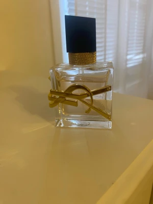 Yves Saint Laurent Libre parfym EDT - Elegant parfymflaska från Yves Saint Laurent med genomskinligt glas, svart fyrkantigt lock och gulddetaljer i form av YSL-loggan. Flaskan har en lyxig och modern design som verkligen sticker ut på hyllan.