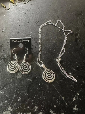 Silverset med spiralhalsband & örhängen - Säljer ett snyggt smyckeset med halsband och matchande örhängen i silverfärgad metall. Både halsbandet och örhängena har en cool spiraldesign som ger en modern och trendig look. Perfekt för att lyfta vilken outfit som helst.