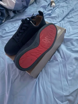 Svarta sneakers med röd sula  - Svarta Stan Smith x Raf Simons sneakers med klassisk låg siluett, svarta skosnören och röd sula med Raf Simons signatur. Ovandel i mocka och detaljer i läder. Perfekta för dig som gillar stilrena och unika sneakers.