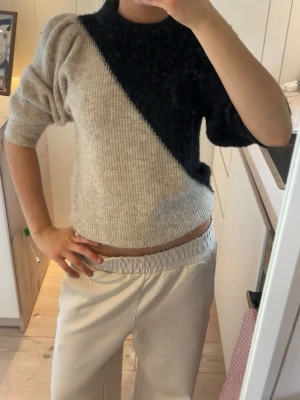 Stickad tröja med svart och beige diagonal - Säljer en cool stickad tröja med diagonal färgdelning i svart och beige. Tröjan har puffiga axlar, halvlånga ärmar och ribbstickade muddar. 