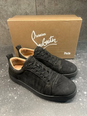 Christian Louboutin svarta sneakers - Svarta sneakers från Christian Louboutin i mocka med klassisk röd sula. Lågt skaft, svarta skosnören och diskret branding. Insidan är ljusbeige och skon har en stilren, minimalistisk design som passar till många outfits.