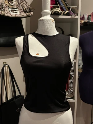 Bik Bok Cut Out Topp  - Svart topp från Bik Bok med trendig asymmetrisk cut out-detalj framtill. Enkel men edgy modell som passar perfekt till jeanskjol, cargobyxor eller lågmidjade jeans för en clean Y2K-look. Materialet har lätt glans/satinliknande finish som ger toppen en lite mer dressad känsla.
