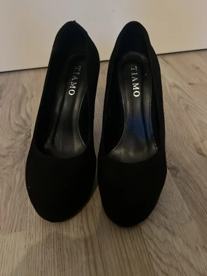 Svarta klackskor från Tiamo - Stilrena svarta pumps från Tiamo med rund tå och hög klack. Skorna är i mockaimitation och har en klassisk siluett som passar till många olika looks. Snygg detalj med Tiamo-logga på sulan. Perfekta för dig som gillar en tidlös och elegant stil.