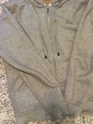 Grå zip-hoodie från Burberry - Snygg grå hoodie med dragkedja från Burberry. Klassisk huva med dragsko och ikoniskt rutigt Burberry-foder på insidan. Mjukt material och ribbade muddar. Perfekt för en chill och stilren look. Det står xl i nacken men den som en small eftersom att det är kinesiska storlekar jag är 172cm lång och den sitter perfekt! Och den är nästan helt ny använd cirka 1-3 ggr