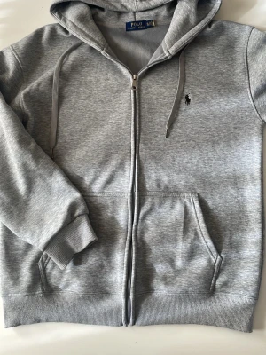 Grå zip hoodie från Polo Ralph Lauren - Snygg grå hoodie från Polo Ralph Lauren med dragkedja, huva med dragsko och två fickor framtill. Klassisk logga broderad på bröstet. Tillverkad i mjuk bomullsblandning och har ribbade muddar vid ärmslut och nederkant. Perfekt för en chill och stilren look. Pris kan diskuteras !!!!