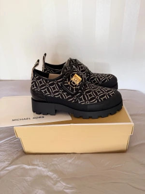 Mönstrade loafers från Michael Kors - Snygga loafers från Michael Kors i svart och grått med grafiskt MK-mönster. Skorna har chunky sula och dekorativt guldfärgat spänne på ovansidan. Tillverkade i textil och syntetmaterial för en modern look.