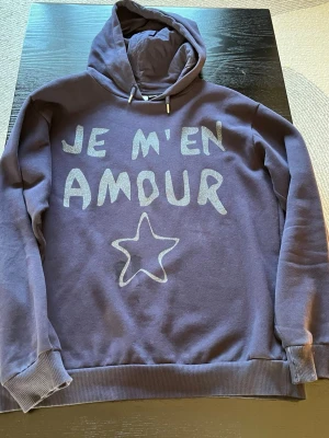 Lila hoodie med tryck från Gina Tricot - Snygg lila hoodie från Gina Tricot med texten 'JE M'EN AMOUR' och en stjärna framtill. Hoodien har dragsko i huvan och ribbade muddar vid ärmslut och nederkant. Perfekt för en chill och trendig look.