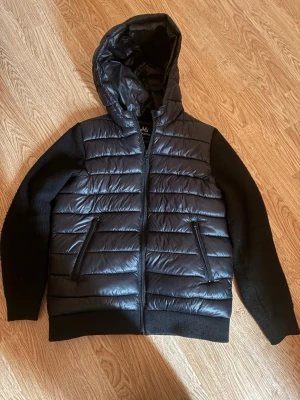 Svart Cardigan från MAGGIORE - Snygg svart pufferjacka från Maggiore med stickade ärmar och vadderad kropp. Jackan har huva, dragkedja framtill och två fickor med dragkedja. Maggiore-logga på ärmen. Perfekt för kalla vinterdagar och riktigt stilren look.