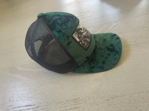 Grön Hatstore  med tryck - Snygg truckerkeps i grön sammet med svart mesh på sidorna och baksidan. Framtill finns ett svartvitt tryck med stadsmotiv. Kepsen har böjd skärm och justerbar passform. Perfekt för dig som vill sticka ut med en unik accessoar.