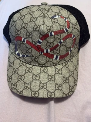 keps med orm  - Snygg keps  med klassiskt GG-monogram i beige och brunt, prydd med en röd, vit och svart orm på framsidan. Baksidan är i svart mesh för extra ventilation. Perfekt accessoar för en trendig streetstyle-look.