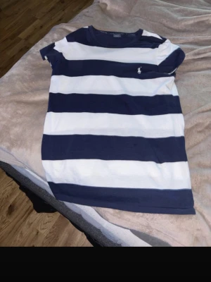 Randig t-shirt från Polo Ralph Lauren - Säljer en snygg t-shirt från Polo Ralph Lauren med breda marinblå och vita ränder. Klassisk passform, kortärmad och med det ikoniska logobroderiet på bröstet. Perfekt för dig som gillar stilrena och sportiga plagg. Liten i storleken