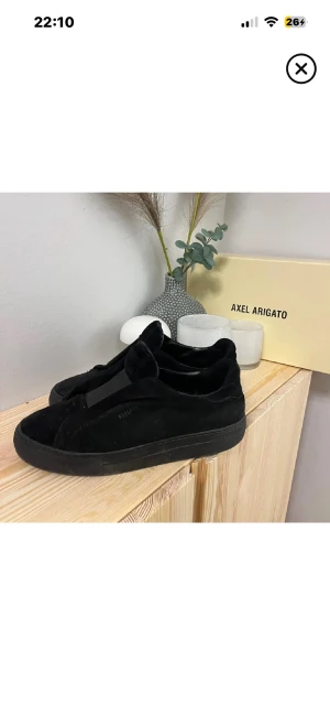 Svarta sneakers från Axel Arigato - Stilrena helsvarta sneakers från Axel Arigato i mocka med låg profil och tjock sula. Skorna har minimalistisk design utan snörning och diskret logga på sidan. Perfekta för dig som gillar en clean och modern look.