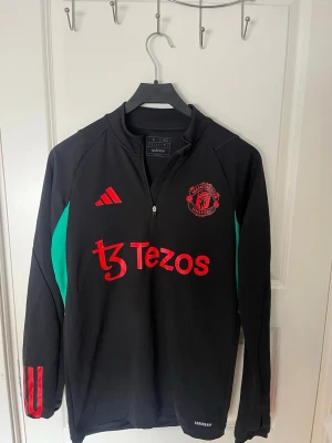 Manchester United träningströja Adidas - Svart långärmad träningströja från Adidas med Manchester United-logga och röd Tezos-sponsor på bröstet. Tröjan har gröna paneler på sidorna, halv dragkedja och röda detaljer på ärmen. Perfekt för fotbollsträning eller sportiga dagar.