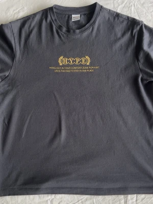Mörkgrå HYPE t-shirt med tryck - Snygg mörkgrå t-shirt från HYPE med gult tryck och text på bröstet. Klassisk rund hals och korta ärmar. Perfekt för dig som gillar streetwear och vill ha en enkel men stilren look.