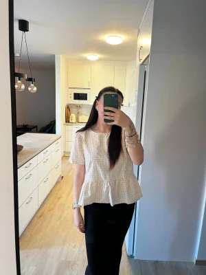 Rutig blus med knyt i ryggen - Supersöt rutig blus i ljusbeige och vitt med korta ärmar och volang nertill. Baksidan har två snygga knyt som ger en unik touch. Helt oanvänd🫶🏼