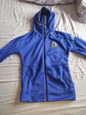 Blå Chelsea hoodie från Adidas - Snygg blå hoodie med dragkedja från Adidas och Chelsea FC-logga på bröstet. Tröjan har huva, två fickor och Adidas-logga på ärmen. Perfekt för dig som älskar fotboll och vill ha en sportig stil.