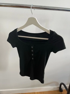 Svart ribbad topp med knappar - En svart ribbad topp med korta ärmar och rundad halsringning. Toppen har fyra knappar framtill som snygg detalj och är figurnära i modellen. Perfekt att matcha med jeans eller kjol för en stilren look.