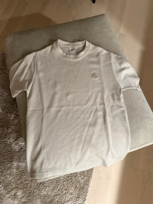 Beige Moncler t-shirt med logga - Snygg beige t-shirt från Moncler med klassisk rund hals och korta ärmar. T-shirten har en stilren broderad Moncler-logga på bröstet och är tillverkad i mjuk bomull. Perfekt för en clean och avslappnad look.