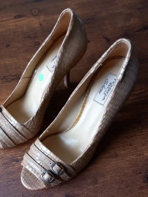 Tosca Blu Shoes Klackskor Beige Strl 37 - Eleganta klackskor med ormskinnsmönster och öppen tå. Skorna har dekorativa spännen framtill och en smal klack. Perfekta för festliga tillfällen eller för att lyfta en vardagsoutfit.