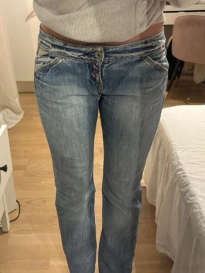 Blå bootcut jeans från Replay - Snygga blå bootcut jeans från Replay med slitna detaljer och låg midja. Jeansen har klassisk femficksdesign, ljus tvätt och synliga sömmar. Perfekta för en avslappnad och trendig look. W28L34