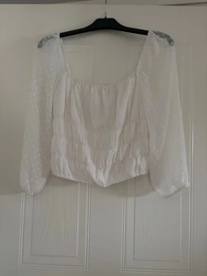 Vit blus med prickiga ärmar - Söt vit blus med transparenta, långärmade ärmar i prickigt mönster. Toppen har en rynkad, luftig passform och bred urringning. Perfekt för dig som gillar romantisk och trendig stil.