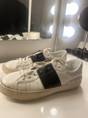 Valentino skor - Snygga vita sneakers från Valentino med en bred svart läderdetalj på sidan. Klassisk låg modell med vita snören och gummisula. Perfekta för en stilren och trendig look.