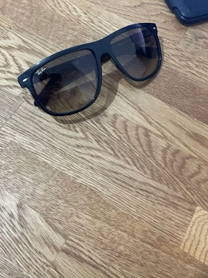 Svarta Ray-Ban solglasögon Wayfarer - Snygga svarta Ray-Ban Wayfarer solglasögon med klassisk form och mörka glas. Ikonisk logga på ena linsen och robust båge i plast. Perfekta för att lyfta din stil under soliga dagar.
