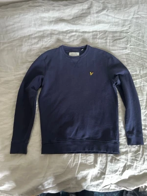 Mörkblå sweatshirt från Lyle & Scott - Mörkblå sweatshirt från Lyle & Scott med rund halsringning och klassisk gul logga broderad på bröstet. Tröjan har långa ärmar, ribbade muddar och är tillverkad i mjuk bomullsblandning. Perfekt för en avslappnad och stilren look.