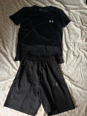 Svart träningsset från Under Armour - Säljer ett svart träningsset från Under Armour med t-shirt och shorts. Båda plaggen har logga på framsidan och är tillverkade i ett lätt och snabbtorkande material, perfekt för gym eller löpning.