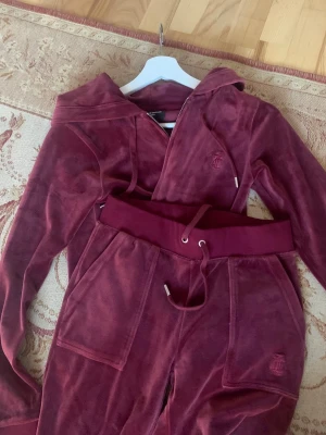 Vinrött velourset från Juicy Couture - Mysigt loungewear-set i vinröd velour från Juicy Couture. Setet består av en hoodie med dragkedja och matchande byxor med bred resår och dragsko i midjan. Båda delarna har broderad logga framtill och stora fickor. Perfekt för chill dagar hemma.