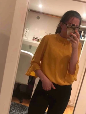 Blus - Senapsgul blus med volanger. Sytt på en vit knapp i nacken, inget som syns när tröjan är på. Samt klippt bort alla lappar 