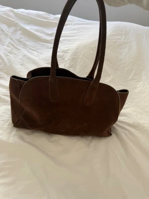 Brun mocka handväska med långa handtag - Snygg handväska i mörkbrun mocka med mjuk, rundad form och långa dubbla handtag. Perfekt storlek för att få plats med allt du behöver. Väskan har en stilren och minimalistisk design som passar till många olika outfits.