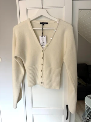 Kofta från GinaTricot XS - Trendig krämvit cardigan från GinaTricot med v-ringning och dekorativa pärlknappar framtill och längs axeln. Modellen har vida ärmar och bred ribbstickad midja som ger en snygg siluett. Helt nytt skick aldrig använt, Ord pis 400kr.