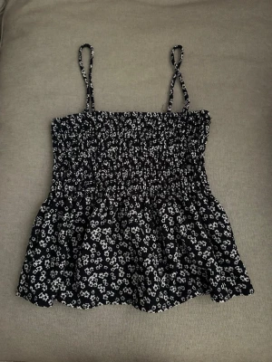 Blommig svart linnetopp Gina Tricot - Supersöt svart linnetopp från Gina Tricot med vita små blommor och smock över bysten. Tunna axelband och lätt utsvängd nertill, perfekt för varma dagar. Toppen har en luftig och bekväm passform som känns både trendig och enkel att styla.
