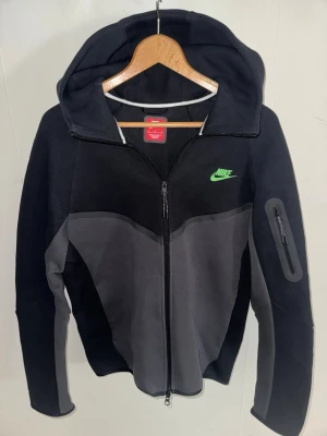Nike Tech Fleece hoodie svart/grå - Nike Tech Fleece hoodie i svart och grått med hel dragkedja och huva. Har en grön Nike-logga på bröstet och en extra ficka med dragkedja på ärmen. Tillverkad i mjukt och lätt fleecematerial som ger en modern look.