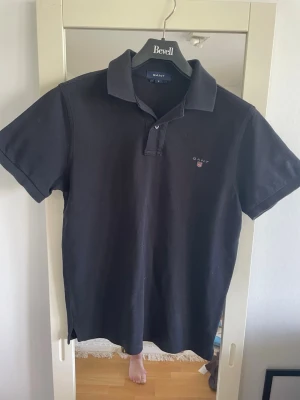 Svart pikétröja från Gant - Klassisk svart pikétröja från Gant med korta ärmar och krage. Tröjan har knappar vid halsen och en liten broderad logga på bröstet. Tillverkad i mjuk bomull som känns skön mot huden. Perfekt för en stilren och avslappnad look.