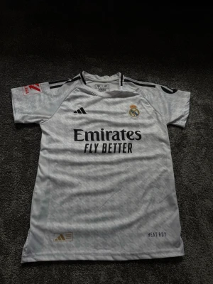 Real Madrid matchtröja Adidas vit - Säljer en vit Real Madrid matchtröja från Adidas med svarta detaljer och klubbmärke på bröstet. Tröjan har trycket 'Emirates Fly Better' på framsidan och är tillverkad i ett lätt, tekniskt material med HEAT.RDY-teknologi. Perfekt för fotbollsträning eller match.