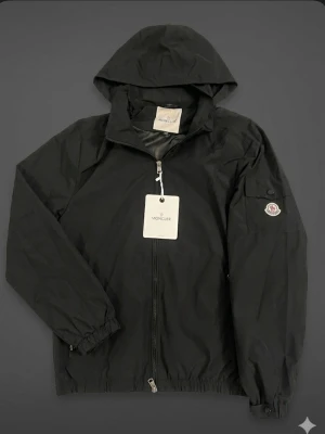 Svart vindjacka från Moncler - Snygg svart vindjacka från Moncler med huva och dragkedja framtill. Jackan har två fickor med dragkedja och en Moncler-logga på ärmen. Tillverkad i lätt och slitstarkt material, perfekt för blåsiga dagar.