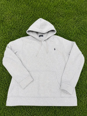 Grå hoodie från Polo Ralph Lauren - Snygg ljusgrå hoodie från Polo Ralph Lauren med klassisk broderad logga på bröstet. Hoodien har huva med dragsko, magficka och ribbade muddar. Perfekt för en chill och stilren look. Materialet är mjukt och skönt, troligtvis bomullsblandning.